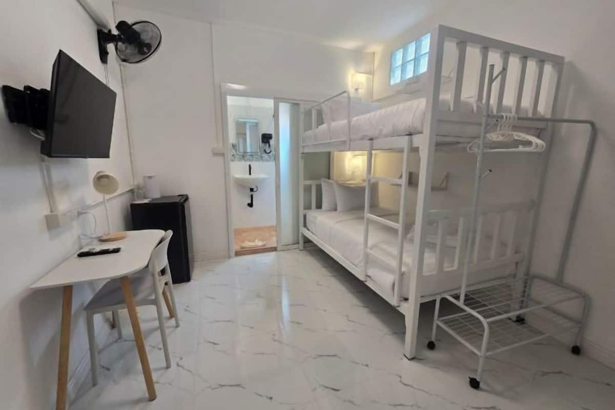 Un lit superposé avec une structure en métal, un bureau avec une chaise, un téléviseur fixé au mur et une salle de bain visible à travers une porte ouverte.