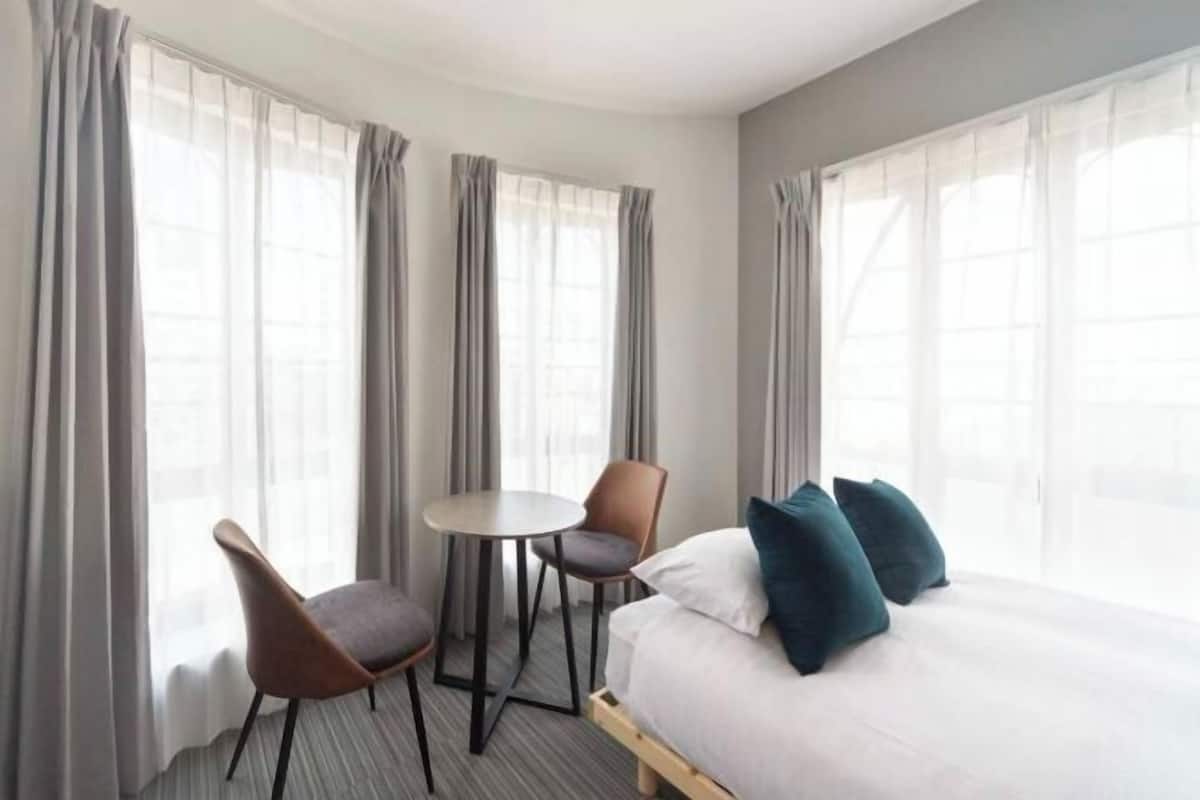Une chambre d’hôtel avec un lit, deux chaises, une petite table et de grandes fenêtres avec des rideaux.
