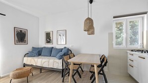 Eine moderne Küche mit Esstisch, Stühlen und einer Couch. An den Wänden hängen Bilder, eine Pendelleuchte und ein Fenster mit Blick auf Grünflächen.