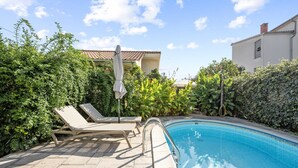 Ein Pool mit Liegestühlen und einem Sonnenschirm, umgeben von Grünflächen und einem Gebäude mit Ziegeldach.