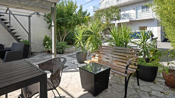 Eine Terrasse im Freien mit einer Holzbank, Korbstühlen und einem Tisch. Übertöpfe mit Pflanzen und eine Treppe, die zu einer erhöhten Ebene führen.