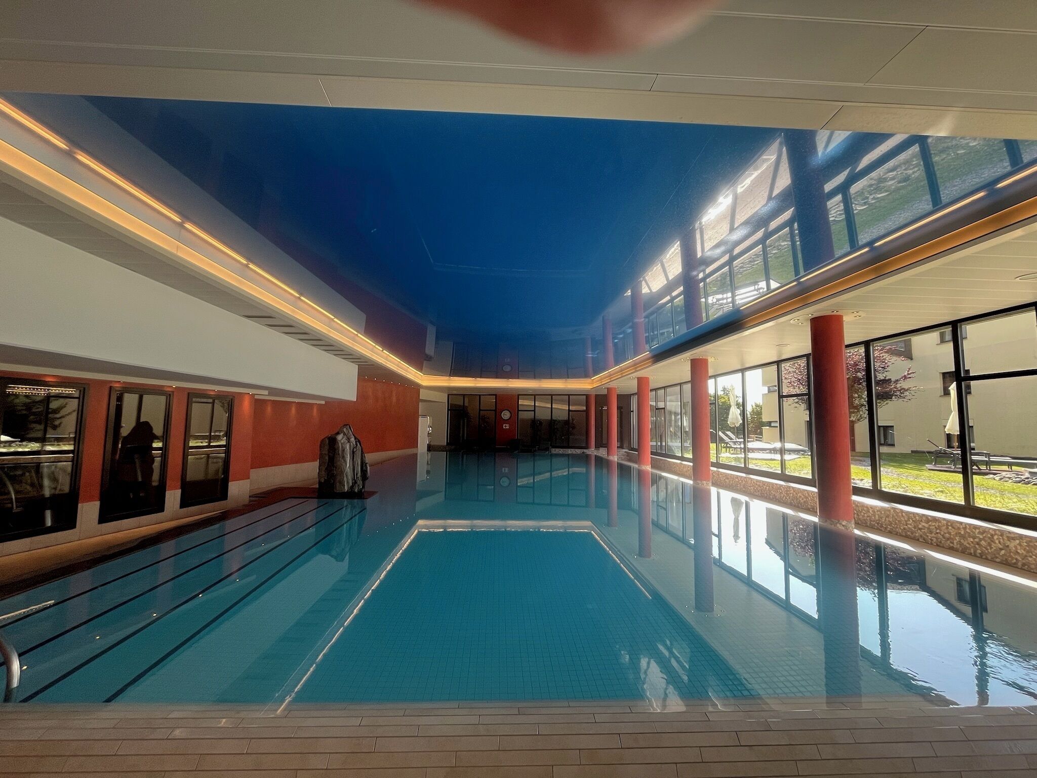 Une piscine intérieure à l’eau d’un bleu cristallin, entourée d’une architecture moderne avec de grandes fenêtres et des piliers rouges.