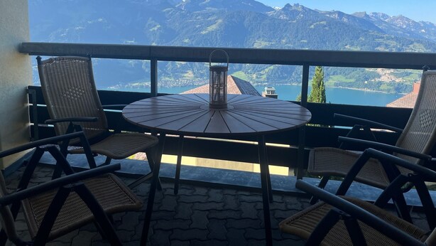 Un balcon agrémenté de fauteuils en osier et d’une table ronde, offrant une vue sur un paysage montagneux et un lac.