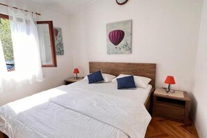 A bedroom with a large bed, two bedside tables with lamps, a window with sheer curtains, and a wall with a framed artwork.