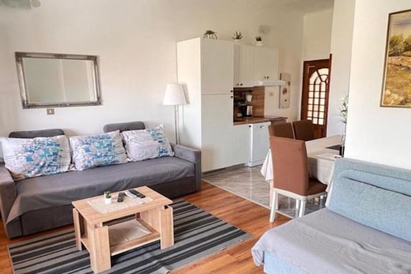 A modern living room with a grey sofa, a wooden coffee table, and a grey armchair. There is a kitchen area with cabinets and a dining table.