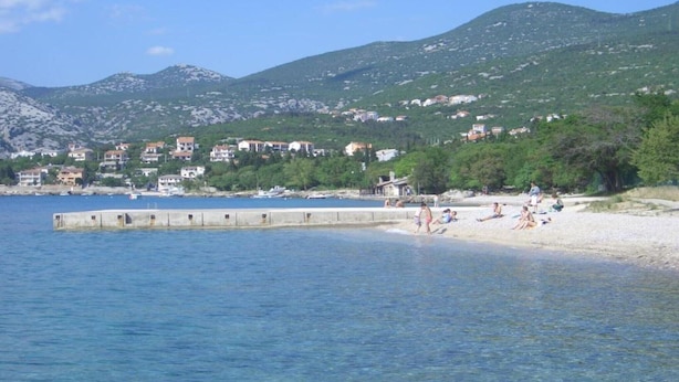Eine Küstenlandschaft mit Sandstrand, einer sich ins Wasser erstreckenden Mole und Gebäuden im Hintergrund.