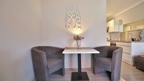 A modern living room with a white table and two grey armchairs, a vase with flowers, and a wall-mounted sign reading 'SEA SIDE'.