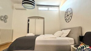 Une chambre d’hôtel moderne équipée d’un lit, d’une table de chevet, d’une chaise, d’une horloge murale et d’un luminaire au plafond.