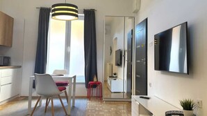 Une cuisine moderne avec des meubles blancs, un coin repas comprenant une table et des chaises blanches, un tabouret rouge et un grand téléviseur fixé au mur.