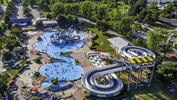 Vista aerea di un parco acquatico con diverse scivoli e piscine, circondato da parcheggi e alberi.