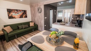 Un salon moderne avec un canapé vert, une table à manger agrémentée d’un centre de table composé de fruits, et un coin cuisine visible à travers une porte ouverte.