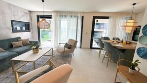 A modern living room with a sofa, coffee table, and chairs, a dining area with a table and chairs, and a kitchen visible through an open door.