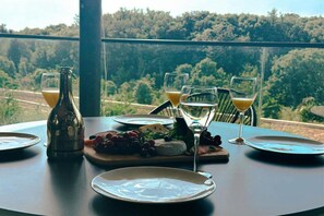 A table set with plates, glasses, a bottle, and a cheese board with grapes and greens, overlooking a forested landscape.
