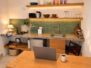 Een kleine keuken met een laptop op tafel, een koffiekop en diverse keukenspullen op het aanrecht.