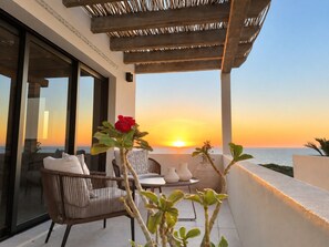 Un balcón con mobiliario de mimbre, una puerta de cristal que da a una habitación y vistas al atardecer.