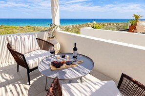 Une terrasse avec une table ronde, des chaises en osier, une bouteille de vin et une vue sur l’océan.