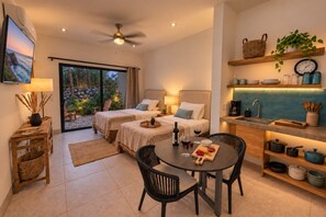 A hotel room with two beds, a dining area with a table and chairs, a kitchen area with a sink and shelves, and a large window with a view of a garden.