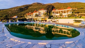 En swimmingpool med solstolar, en byggnad med flera balkonger och en bergig bakgrund.