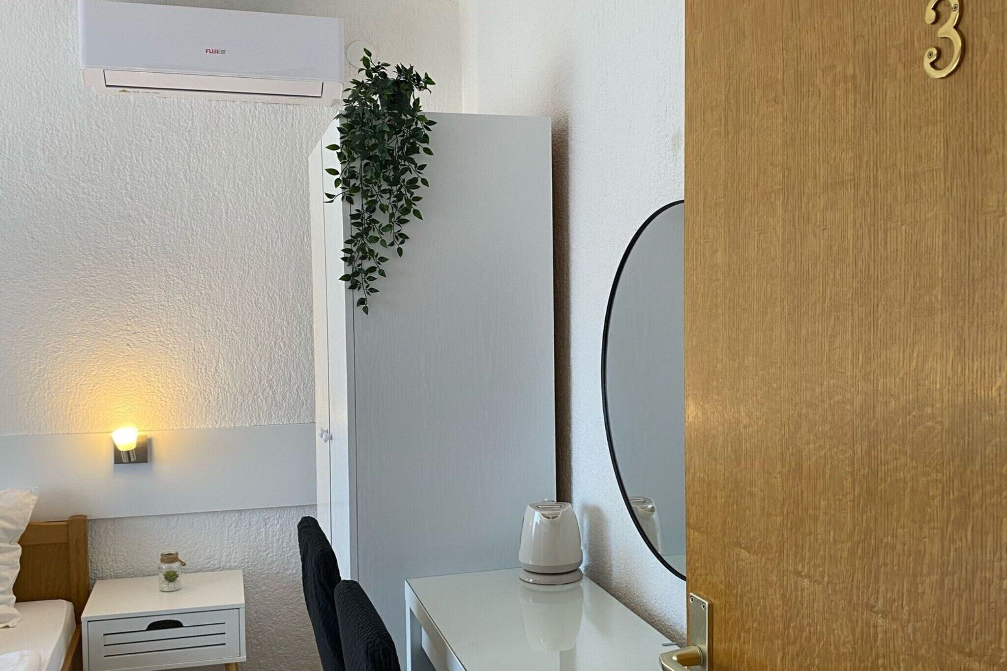 Et hotelværelse med en hvid seng, et sengebord med en lampe, en lille plante, et spejl og en trædør med et nummer.