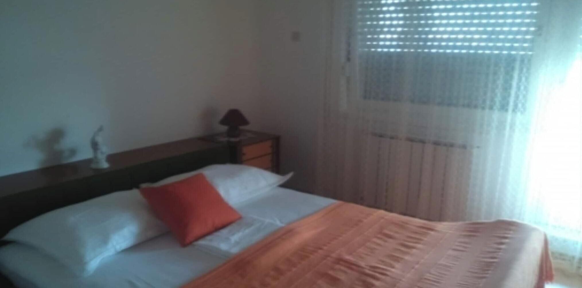 Een slaapkamer met een bed, nachtkastje, lamp en raam met dunne gordijnen.