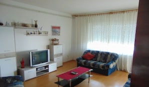 A living room with a television, sofa, and coffee table. There are shelves with decorative items and a window with curtains.