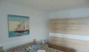 A bedroom with a bed, a bedside table, a nightstand, a painting of a sailboat, and a wooden wardrobe.