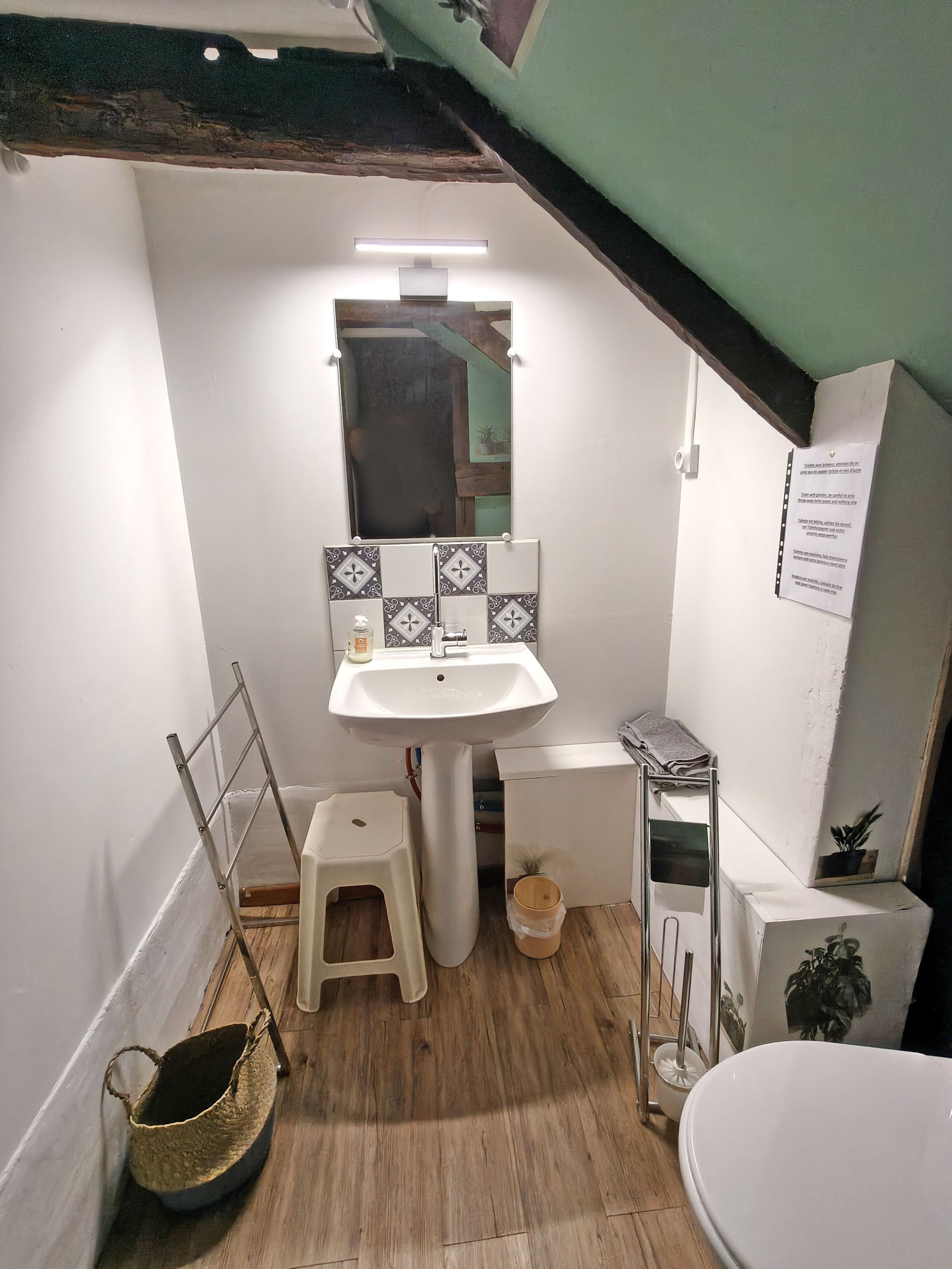 Une petite salle de bain avec un lavabo blanc, un miroir et des toilettes. On y trouve un petit tabouret et un panier posé sur le sol.