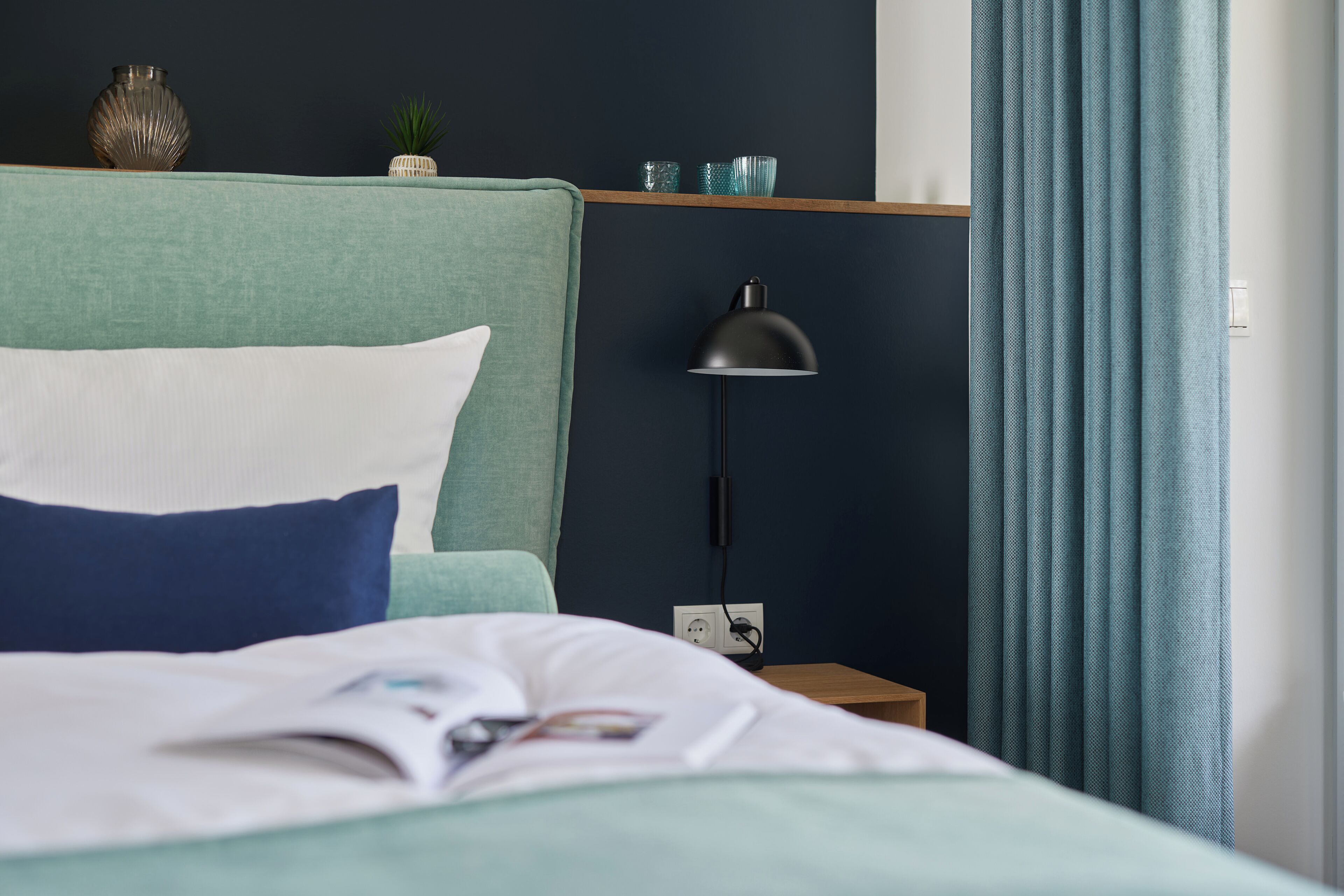 A neatly made bed with pillows, a bedside lamp, a small nightstand with a power outlet, and a shelf with decorative items.