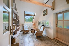 A cozy room with a sloped ceiling, built-in bookshelves, wicker furniture, a small table, and a door with frosted glass panels. - Holiday Home 'Das Künstlerhaus – Your Summer Retreat' with Private Terrace, Garden & Wi-Fi (Köckern)