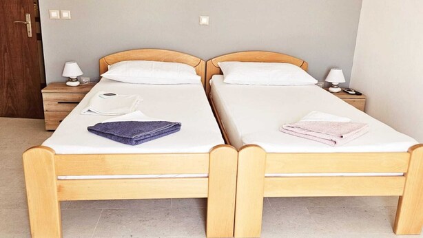 Een kamer met twee houten bedden, elk voorzien van een wit matras en kussen, een nachtkastje van hout met een lamp, en een wandlamp.