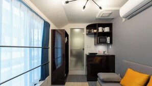 A compact living space with a kitchenette, a sofa with an orange cushion, a TV, and a window with curtains. - LaVie Yotsuya (Tokyo)