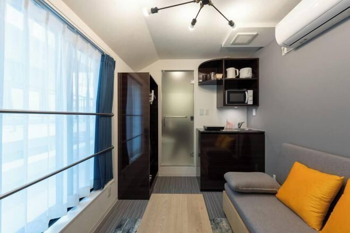 A compact living space with a kitchenette, a sofa with an orange cushion, a TV, and a window with curtains.