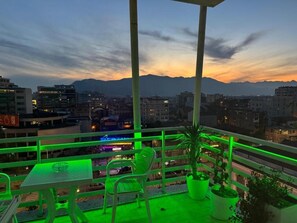 Un balcón con una mesa y sillas, plantas en macetas y vistas a un paisaje urbano con montañas al fondo.