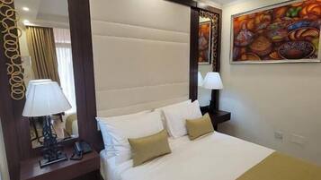 A hotel room with a large bed, a wooden floor, a mirror with a decorative frame, a nightstand with a lamp, and a painting on the wall.