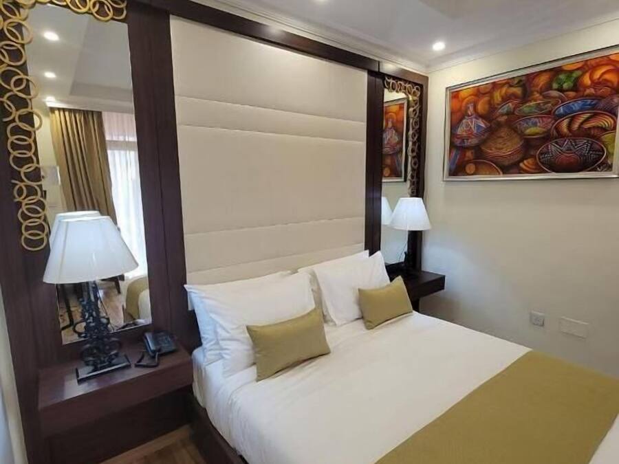 A hotel room with a large bed, a wooden floor, a mirror with a decorative frame, a nightstand with a lamp, and a painting on the wall.