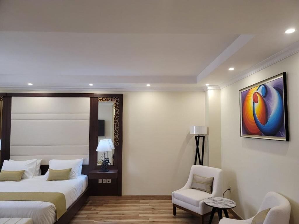 A hotel room with two beds, a sitting area with a chair and a small table, a lamp, and a painting on the wall.