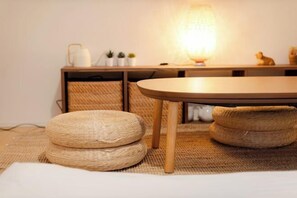 Une pièce avec une table ronde en bois, des poufs en fibres tressées, une lampe et un petit meuble contenant des objets décoratifs.