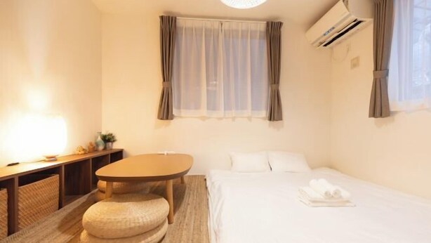 A hotel room with a bed, a round table, a small stool, a bedside table, and a window with curtains.