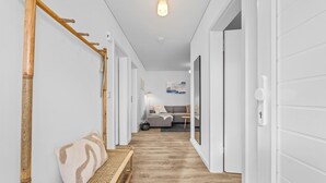 Un couloir doté d’un plancher en bois, d’un banc avec un coussin à motifs et d’une porte donnant sur une pièce où se trouvent un canapé gris et un tableau accroché au mur.