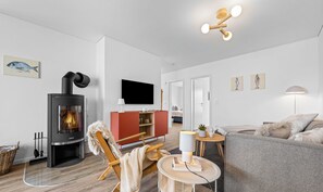 A modern living room with a wood-burning stove, a flat-screen TV, a sofa, a chair, and a small table with a lamp.