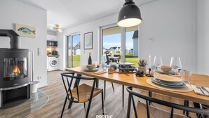 A modern kitchen with a wood-burning stove, dining area with a wooden table and chairs, and a view of houses through large windows.