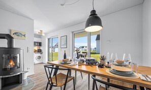 Cozinha moderna com lareira a lenha, área de jantar com mesa e cadeiras de madeira, e vista para casas através de grandes janelas.