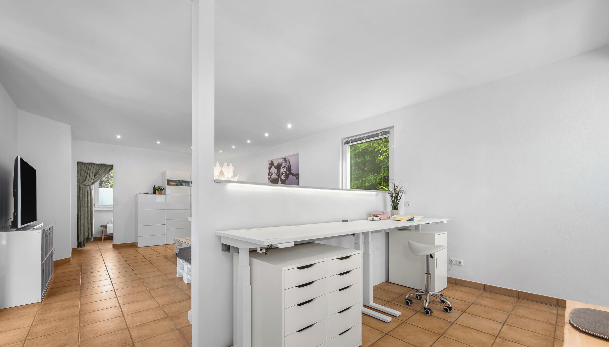 A modern, well-lit room with a white desk, drawers, and a small stool. There is a television on the left and a window with a view of greenery.