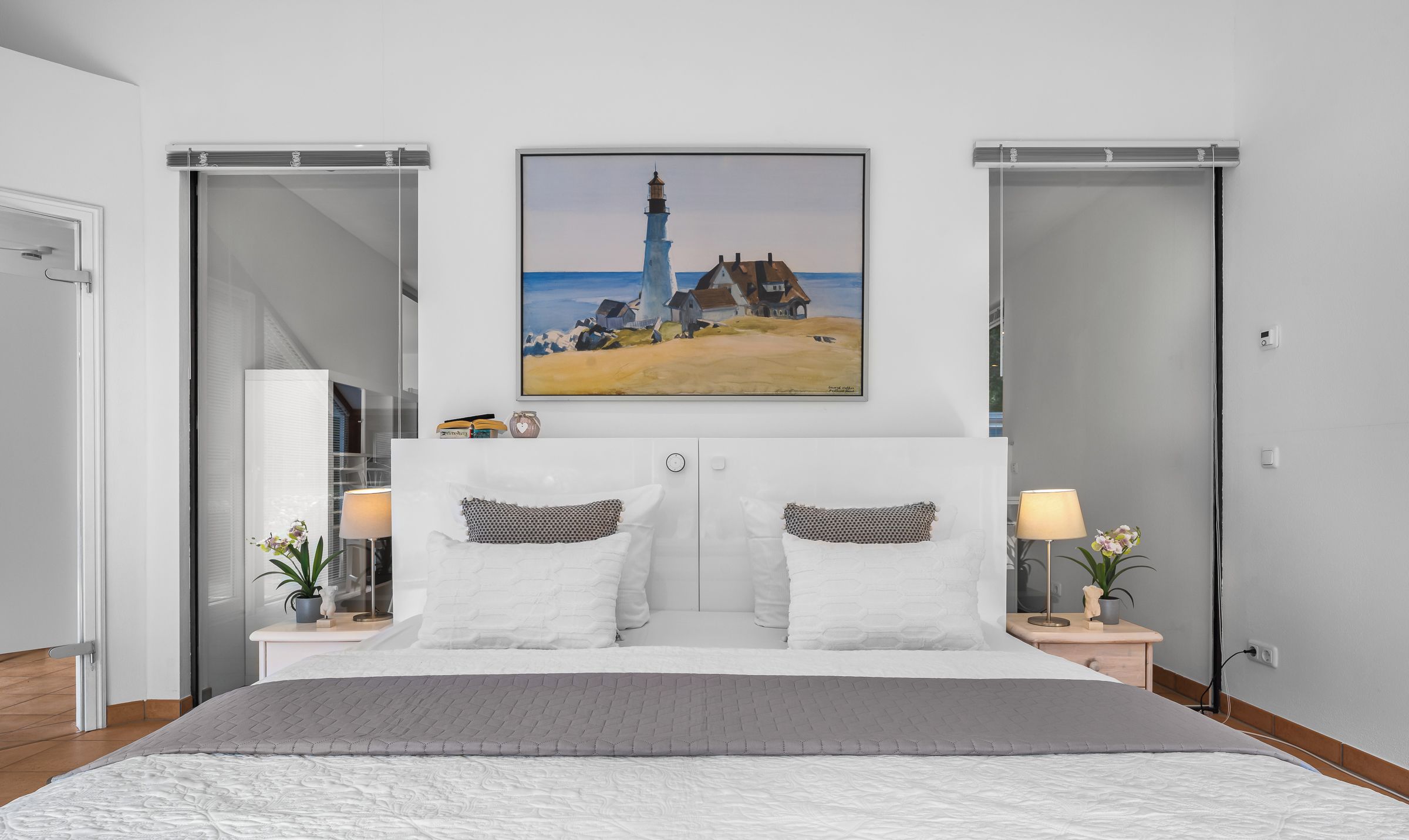 Ein modernes Schlafzimmer mit einem großen Bett, zwei Nachttischen mit Lampen, einem Gemälde eines Leuchtturms und einer Schiebetür.