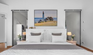Un dormitorio moderno con una cama grande, dos mesitas de noche con lámparas, un cuadro de un faro y una puerta corrediza.