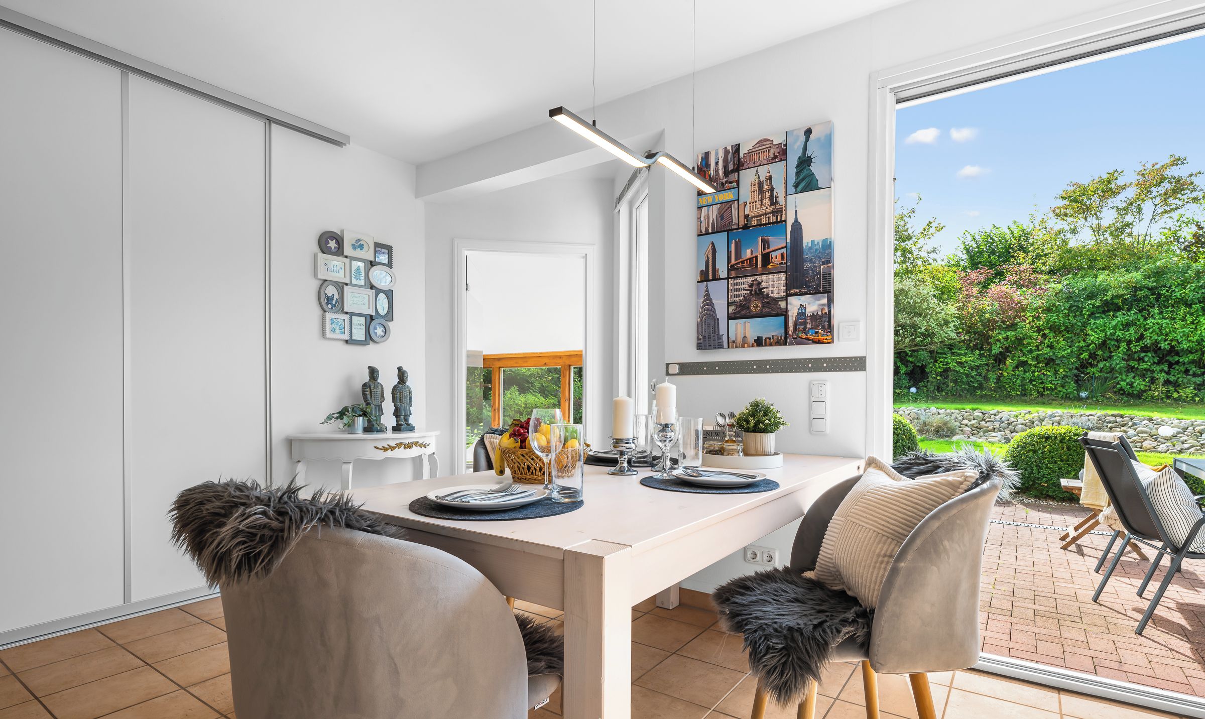 Ein moderner Essbereich mit einem weißen Tisch, zwei Stühlen mit Fellkissen, einer Pendelleuchte und einem Stadtbild-Wandgemälde.