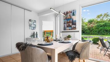 Ein moderner Essbereich mit einem weißen Tisch, zwei Stühlen mit Fellkissen, einer Pendelleuchte und einem Stadtbild-Wandgemälde.