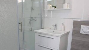 Een moderne badkamer met een douchecabine van glas, een wit wastafelmeubel met een spiegel erboven en een klein plankje met toiletartikelen.