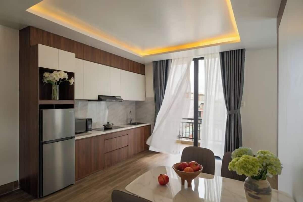 A modern kitchen with a refrigerator, microwave, and wooden cabinets. There is a dining table with a vase of flowers and a bowl of fruit.
