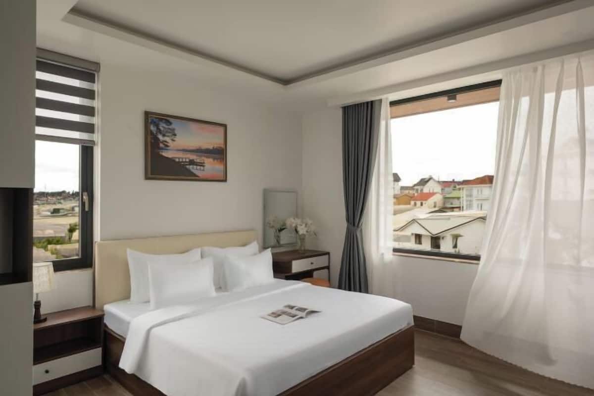 A hotel room with a bed, a bedside table, a window with curtains, a painting on the wall, and a view of buildings outside.
